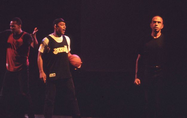 Performance art. Two men with dark skin tones standing to the left wearing basketball jerseys, one holding his arms at 90 degrees, the other a textured orange ball in left hand. Another (light skin tone) man stands half in shadow, clenched hand, wearing a black t-shirt and trousers in this onstage colour photograph.