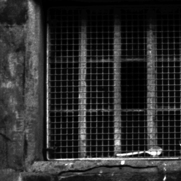Black and white photograph. Stone wall with metal barred window in the majority of this composition. A shiny silver metal spoon is wedged between the window and the stone grid behind, against a dark background.