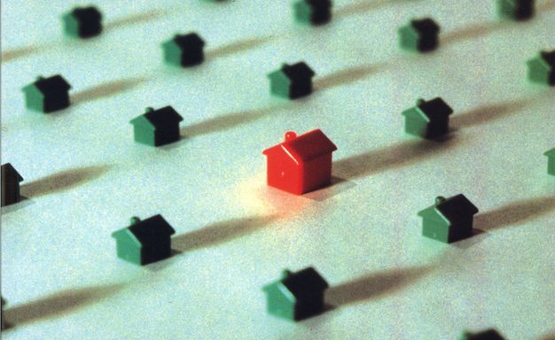 Colour photograph. One red plastic house toy is seen slightly off-centre with several smaller green plastic houses scattered around it. The background of this grainy image is pale green and cream.