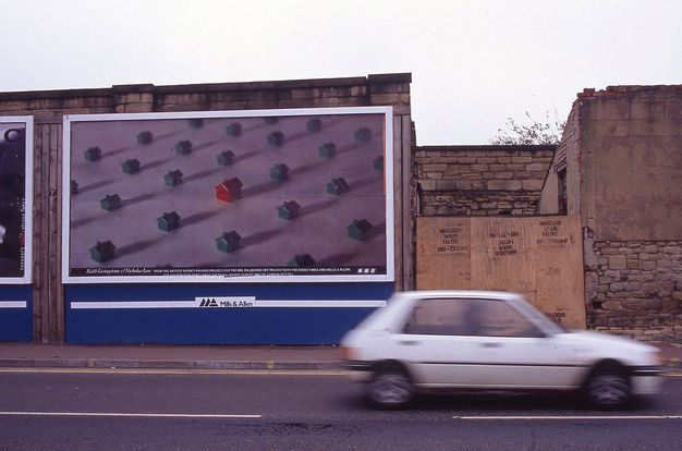 Colour photograph. Street view of stone wall with blurred cream car moving across the foreground. Billboard showing photograph of a red toy house surrounded by smaller green plastic houses besides boarded up wall with repeated spraypainted "WADDS WAS HERE" and telephone number graffiti, shown against a grey sky.