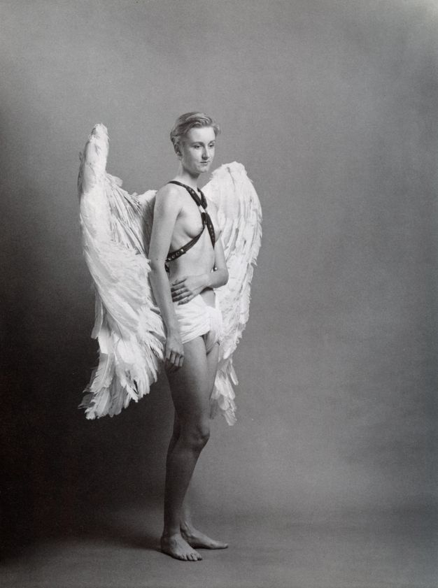 Black and white photograph. Person with combed back light hair stands off-centre, right knee slightly bent, holding their ribs with left hand at a right angle. Wearing a studded leather harness that crosses at their chest and large feathered wings set against a soft textured photography background.