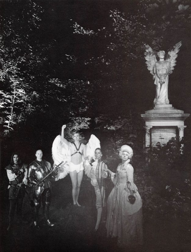 Black and white photograph. Five people standing in a woodland, arranged as orthogonals, converging at person wearing wings on a black leather studded harness with their right arm up. Two people in armour on the left, two people wearing French aristocratic-style clothing on the right and an angel statue in the right of middle ground.