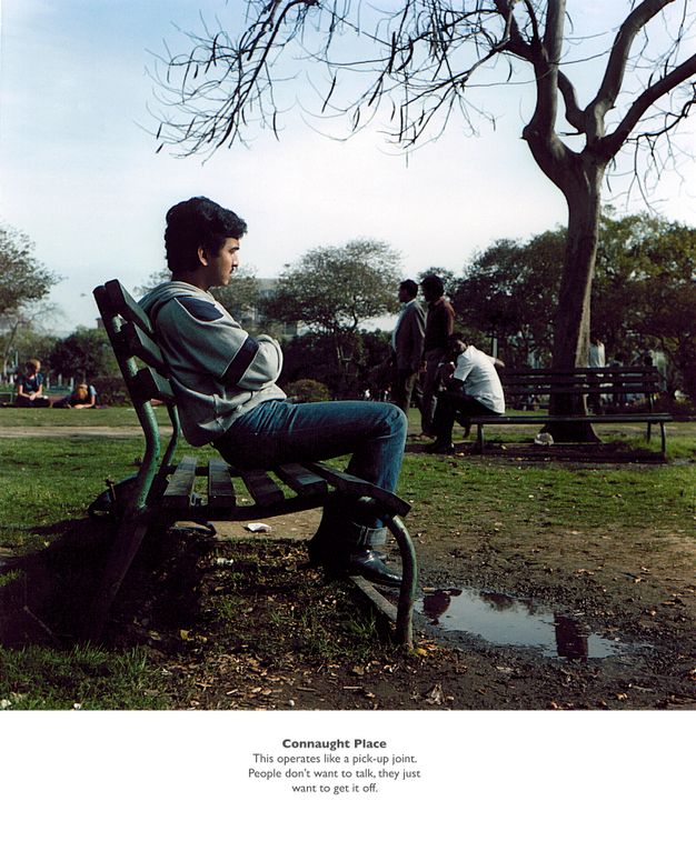 Colour photograph. Man in the central foreground sitting on a green park bench. In the middle ground, three men gather at a second bench by a tree, one looking towards the first man. A horizontal white panel of centred typewritten text "Connaught Place. This operates like a pick-up joint" extends across the lower edge of the composition.