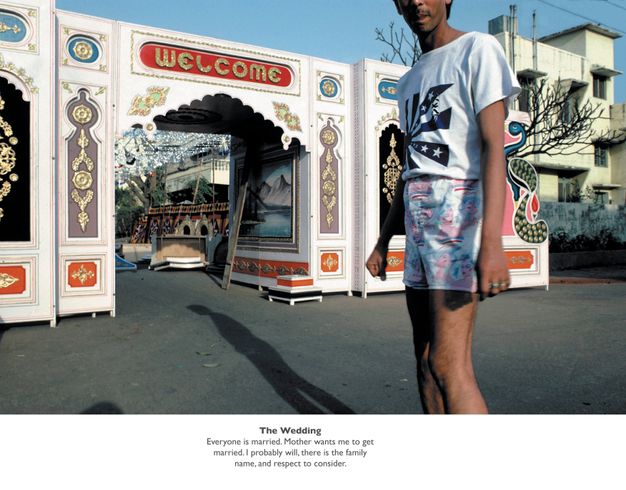 Colour photograph. Man with medium-brown skin wearing pastel pink and green denim shorts and t-shirt stands in front of an ornate wooden gateway decorated in gold and bright colours. Typewritten text including ""The Wedding"" spans the lower quarter of the page on a white background."