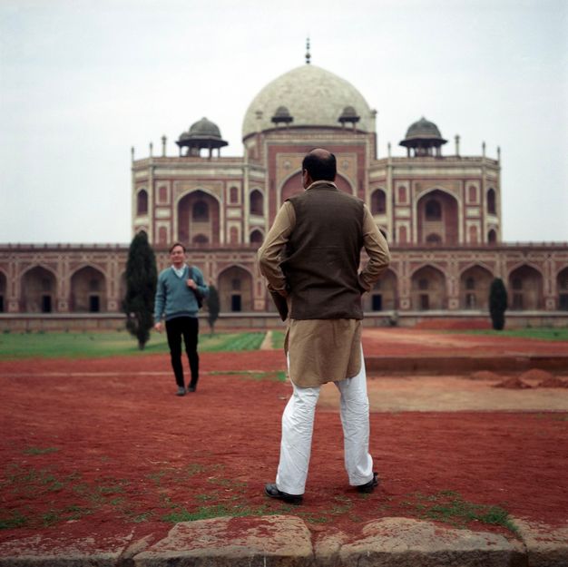 Colour photograph. Man with medium-brown skin wearing white trousers and brown tunic stands on red dusty ground, his back to the camera. A younger man walks in his direction wearing Western clothes with an ornately arched red and cream domed building in the background.