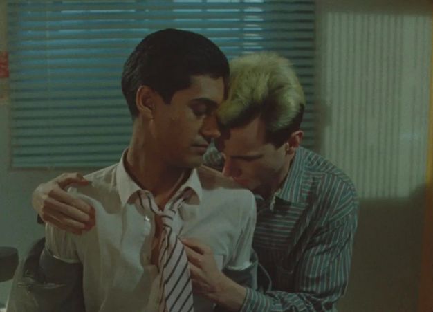 Colour film still. Two men wearing striped cotton shirts and a tie, one places his hands on the second man's shoulder and inside his half-unbuttoned shirt while the latter closes his eyes. Shown in an office setting with slatted window blinds.