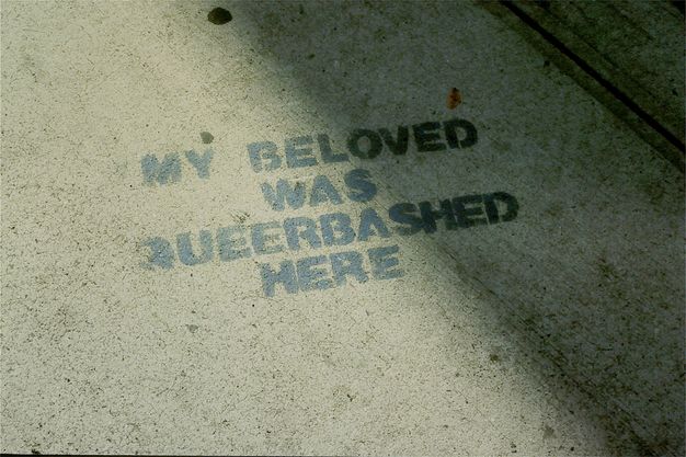 Colour photograph. Textured concrete floor half-covered in shadow and sunlight, with the capitalised grey stencilled text "MY BELOVED WAS QUEER BASHED HERE". The floor has mottled textures and two parallel lines seen in the top-right corner.