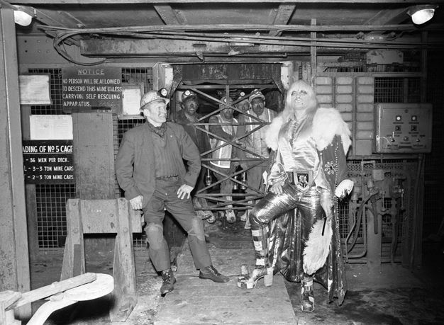 Black and white photograph. A man wearing a miner's hat stands beside a person wearing a wig, patchwork platform boots and satin clothes. Standing behind them are four male miners standing inside a lift shaft with criss-crossed metal gate.