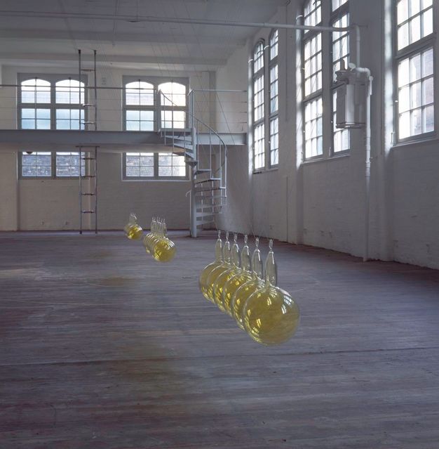 Installation. Three sets of receding six golden glass orbs (with neck sections) suspended from the ceiling of an industrial white warehouse. White metal spiral staircase and mezzanine platform seen in the background with tall, gridded windows.