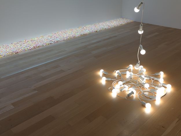 Installation. A string of globe lights in white casings pool on a pale wooden floor with a length suspended above the view of this interior space. A channel of irridescent pebbles arranged along one wall intersecting with another wall, in this corner view of a room.