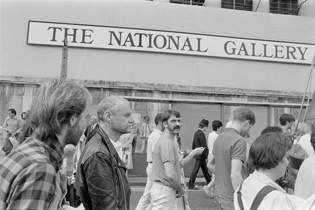 Black and white photograph. A crowd of people with light skin tones walk from left to right of shot past a sign with capitalised text for "THE NATIONAL GALLERY" on a nearby wall. A policeman walks in the same direction, seen in the background of this street scene.