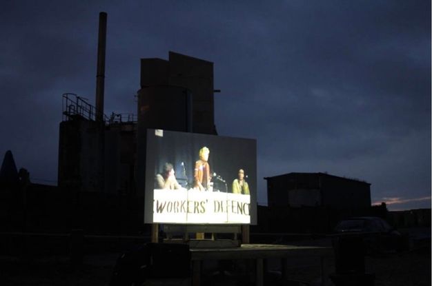 Installation. Film projection in outside space at night with industrial buildings in silhouette. Three figures stand in front of a banner that reads \"WORKERS' DEFENCE\"
