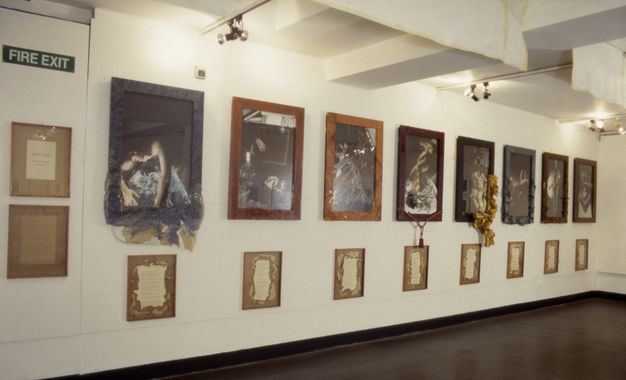 Installation. Eight wooden framed photographs of women span a white wall, three of which have material or a rope tassel descending from the frames. Surrounding these are 10 framed buff and cream parchment panels with typewritten text and ripped borders in a lit interior space.