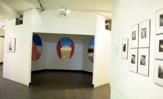 Installation. Eight framed photographs arranged on a white wall on the right side. Hexagonal central exhibition pod showing gold, red and blue collages occupies the middle ground of a white interior space. Metal ceiling spotlights illuminate the space.