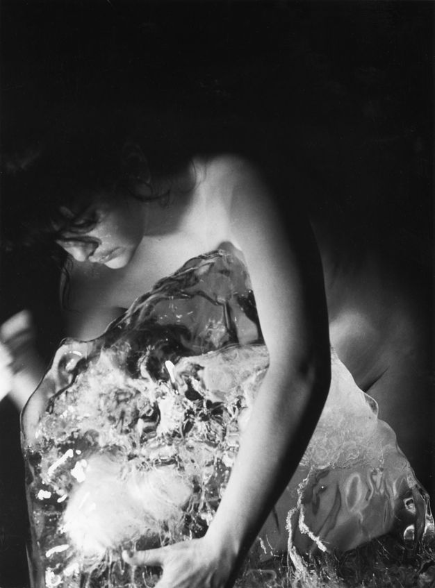 Black and white photograph. Naked woman with curly fronds of hair across her forehead curves her back and leans towards the left, her right hand is blurred while her left hand is placed on a partially jagged semi-transparent object. Solid black background.