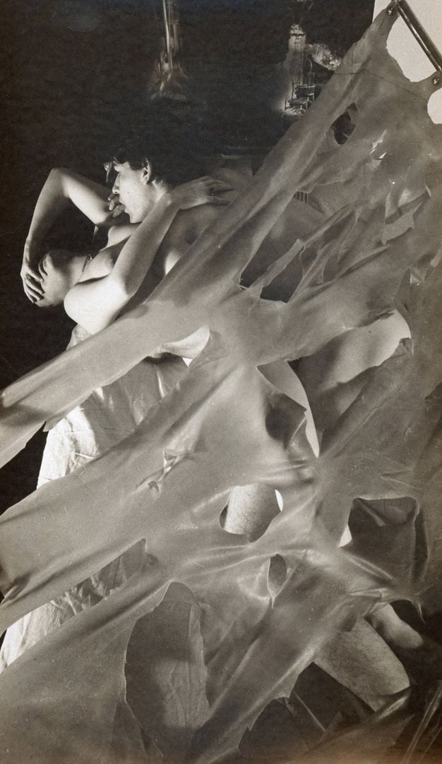Black and white photograph. Layers of thin, ripped sheeting hang diagonally from a metal pole in the upper-right corner, partially covering two intertwined people. The woman has her eyes closed, placing one hand on her face at a 90-degree angle, her other on the man's shoulder. Dark background.