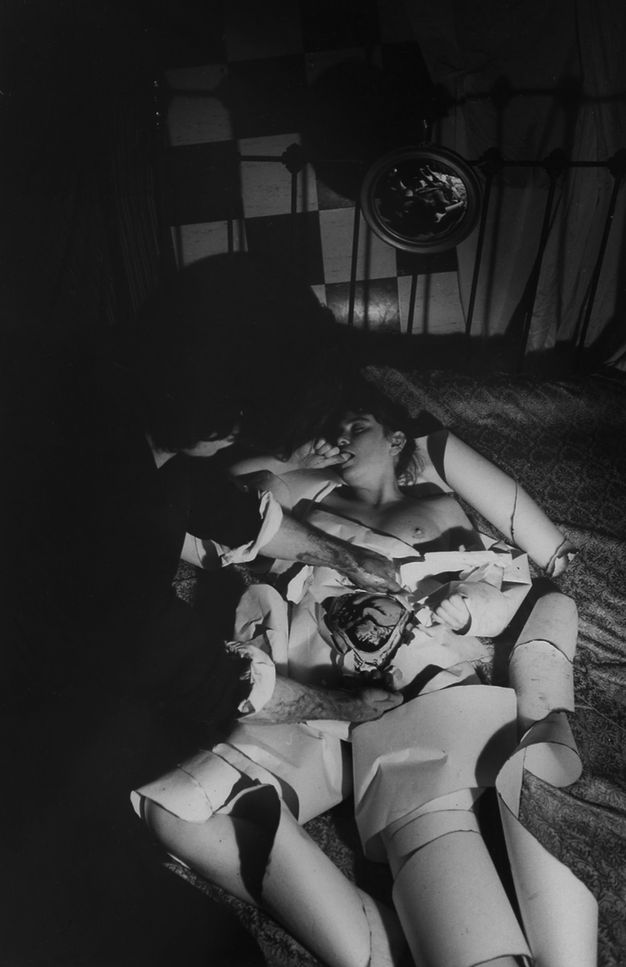 Black and white photograph. Naked woman is lying on a jacquard bedsheet; her abdomen opened revealing a baby in the foetal position. Curled paper wraps around her limbs while a woman wearing dark clothes has her blood covered arm on the foetus. Background includes a metal bedframe and checkerboard wall.