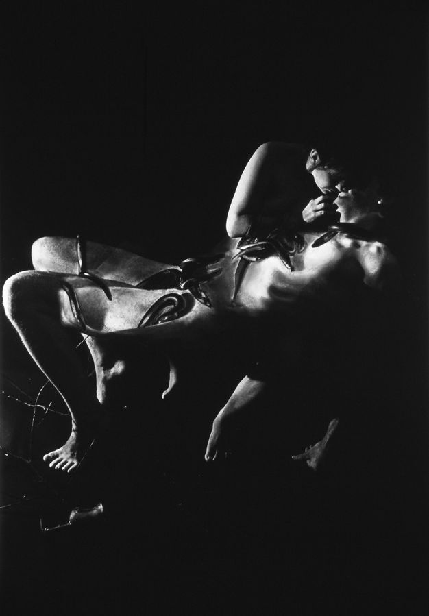 Black and white photograph. Two naked women, one woman's eel-covered body reclines suspended in mid-air revealing her ribcage under the skin, the other woman leans over to her mouth, her leg visible underneath the other's back, near a wire grid in the lower-left corner of the dark background.