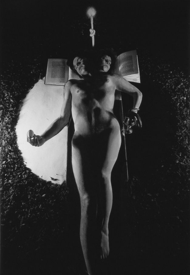 Black and white photograph. A naked person reclines - or is suspended by a small rope - across the centre of the image. They have two heads, each of which rests on an open book. Their body appears split into two, with a masculine, and a feminine, half. The hand on the left holds an egg over a semi-circle of light, while the right holds a wine glass over a semi-circle of darkness.