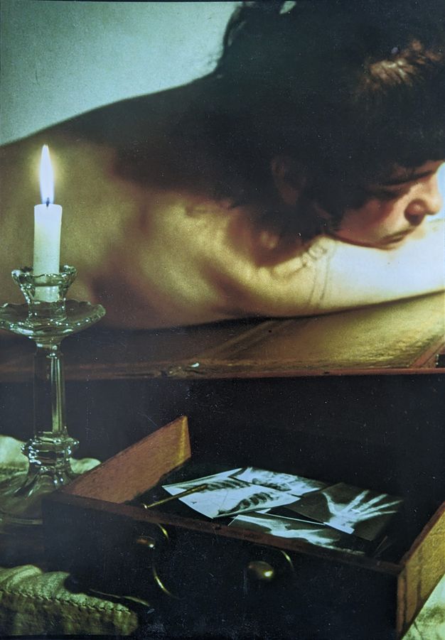 Colour photograph. The shoulders and head of a naked person lying face down across the surface of an antique, fold-out desk. In front of them are a lit candle in a clear glass holder, and an open desk drawer containing a series of bodily x-rays.
