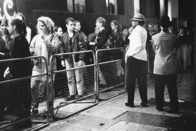 Performance art. In this black and white photograph young adults form a queue behind a metal barricade, seen from a side on view. In the upper left the partial view of a neon sign reveals the letter ‘C’. On the other side of the barricade are two people dressed in white jackets and black trousers. One wears a white hat, and the other a black one.