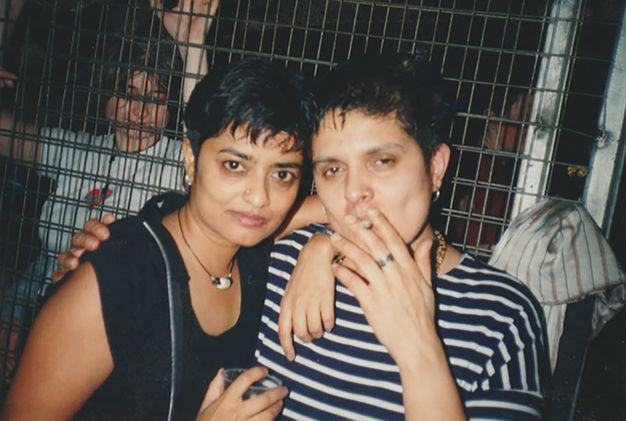 Colour photograph. Two people from the shoulders up face the camera with their arms around one another. One is smoking a cigarette while the other holds a partially seen plastic cup. Behind them is a metal barricade, through which we can see another person in the background.