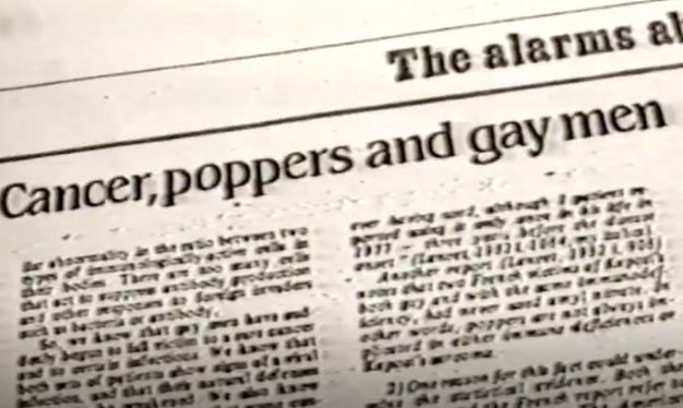 Black and white photograph/video still. In this black and white still from a video, we see a newspaper excerpt at an oblique angle. The visible fragment of article title reads, “Cancer, poppers and gay men.”