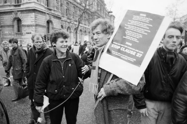 Black and white photograph. Actor Ian McKellan is seen on a crowded street holding a large placard reading, “Stop Clause 27.” A smiling woman stands next to him holding a microphone.