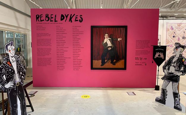 Installation photograph. A freestanding pink wall is emblazoned with text in a hand-drawn style reading ‘REBEL DYKES”. Below the text is a longer passage of text, accompanied by an artwork featuring a person dressed in a black suit against a red curtain. Two freestanding, life-size cut-out illustrations of people dressed in black leather are posed in the space.