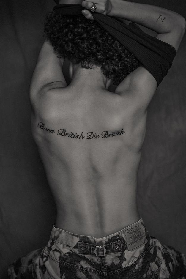 Black and white photograph. A seated person is photographed from behind while removing a top to reveal a tattoo across their upper back reading “Born British Die British” in cursive text. The camera has cropped them at the lower hips, leaving their patterned trousers just visible. On one of their raised, upper forearms another tattoo reads, “Ali + Paul”.
