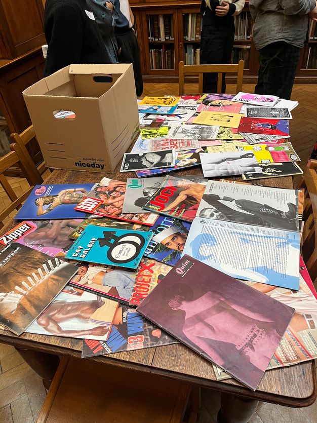 Photograph. A collection of colourful booklets and magazines are spread out across a brown wood tabletop. Magazine titles include “Jock”, and “Zipper”. Also on the table is a brown cardboard box containing more material. The table has wood chairs pushed in against it, and three people stand beyond it.