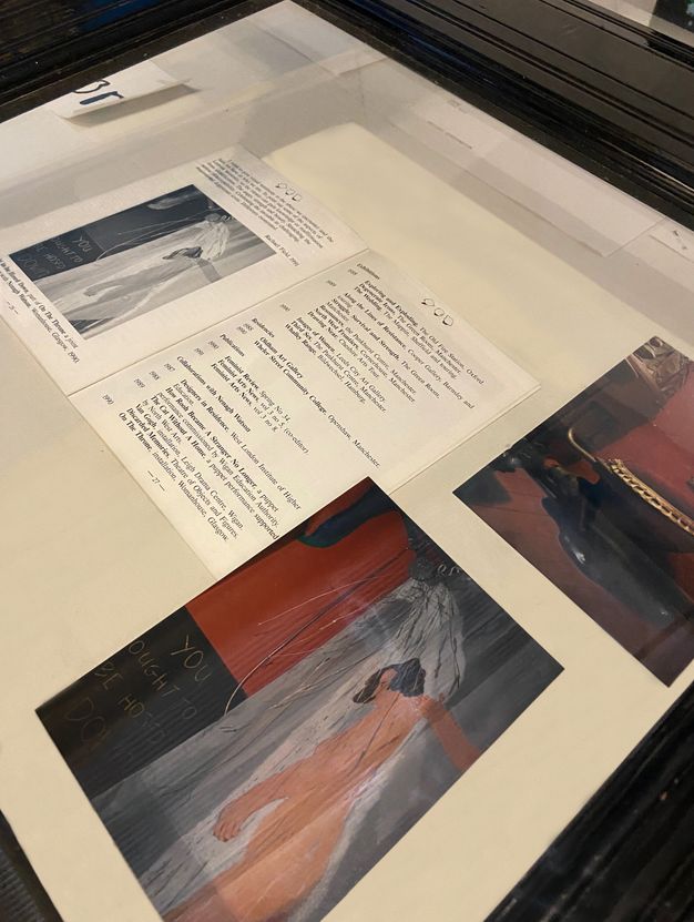 Installation photograph. A display case with a clear glass top contains an open booklet, and two colour reproductions of artworks. In one artwork a naked person stands in a shower, with a red and black wall behind them, and the text “you ought to be hosed down”. The other image is partially cropped but elements of a domestic interior are visible.