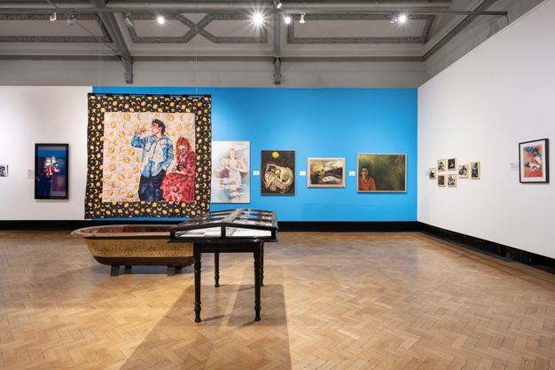 Installation photograph. A gallery with white and blue walls has a series of artworks on display. Hung on the walls are a series of framed and unframed artworks in different styles, while a colourful quilt is suspended from the ceiling. On the floor is a brown and yellow wooden bathtub, and a wooden display case beside it.