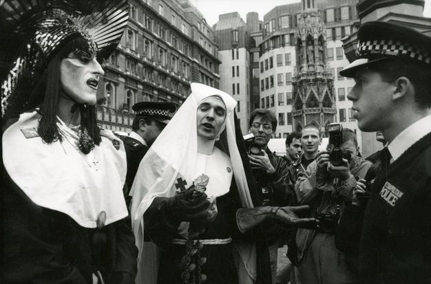 Black and white photograph. Two people wearing nun costumes stand before a policeman on a busy city street. One is wearing drag make-up and an elaborate headdress, and the other heavy leather gloves. A crowd of people gather behind them, one of whom is taking a photograph, while another carries a camera. City buildings are seen in the street beyond.