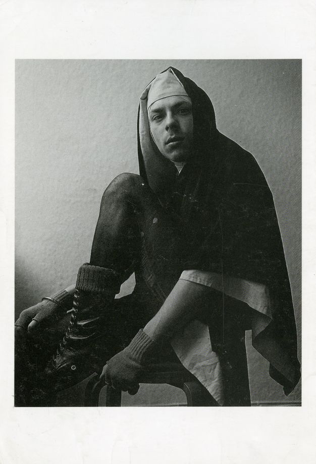 Black and white photograph. A person wearing a nun’s veil and wool, fingerless gloves is seated on a stool, looking out towards the camera against a plain white backdrop. One leg is bent, with a foot perched on the edge of the stool, revealing their thin, laddered tights, thick socks, and black, leather lace-up boots.