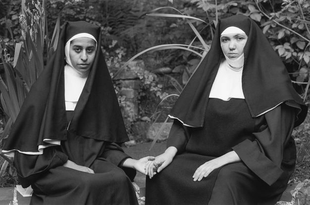 Black and white photograph. Two women wearing traditional nuns’ habits are seated side-by-side on an outdoor stone wall within a secluded, private garden. Holding hands, they look out to the camera, meeting our gaze, while one nun presses her hand between her thighs. Behind them is a garden full of thriving plants.