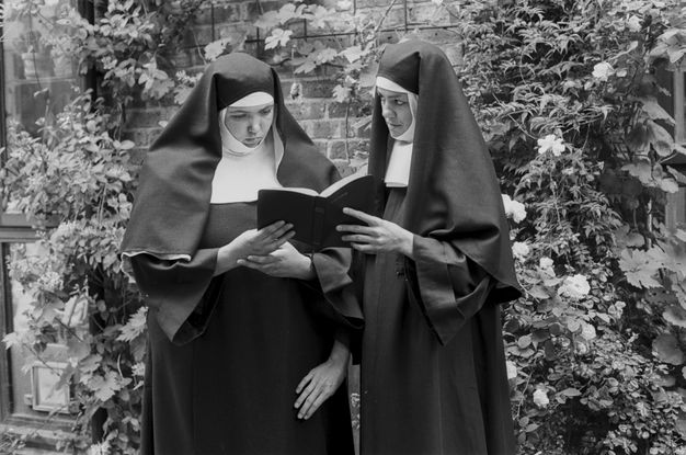Page of a book. Two women wearing full nuns’ habits stand side by side in a secluded garden, holding a bible together. The nun on the right of the image looks suggestively at her companion, while gingerly touching her. The nun on the right stares down at the bible. The image is surrounded by a white frame, with the word “TRANSGRESSION” printed on the upper right, and the word “DEVOTION” on the lower right.