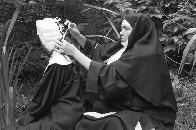 Black and white photograph. Two nuns sit on the ground side by side, surrounded by grass and plants. One is wearing the full nuns’ habit and turns to the side to lace or unlace the partially removed veil from her companion, whose head is turned away from us.
