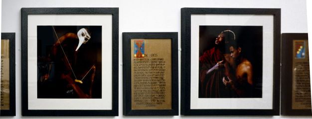 Installation photograph. Five framed artworks hang closely together on a wall. The two on either side are cropped, while the three in the centre can be seen clearly. Two are colour photographs featuring people with a dark skin tone in theatrical costume, while the third is an illuminated text.