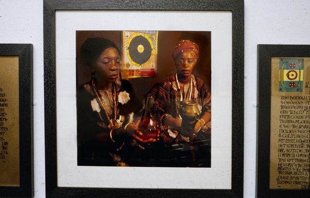 Installation photograph. Three framed artworks sit closely together on a white gallery wall. The two on either side are cropped, but passages of illuminated text can just be seen. The image in the centre is a colour photograph featuring two women with a dark skin tone dressed in colourful clothing, one of whom holds a clear bottle of red liquid in two hands.