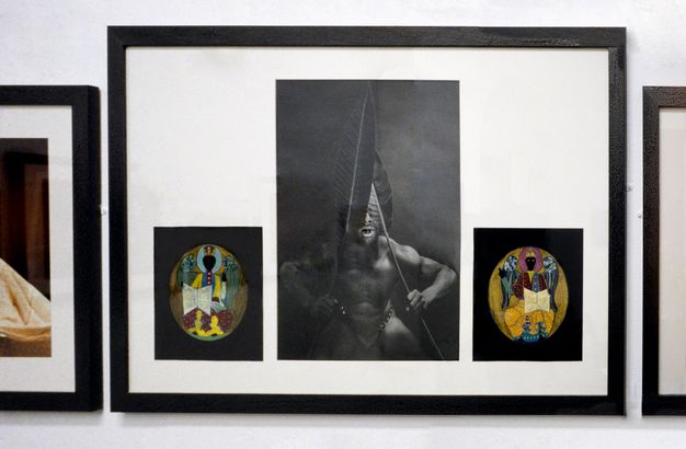 Installation photograph. Three framed artworks are hung closely together. The two on either side are cropped from view. The artwork in the centre is in three parts; a tall, central black and white photograph documents a dark toned man holding two large leaves over his face, while the smaller, flanking images are two colourful, oval designs.