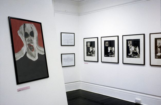 Installation photograph. A view into a gallery space with white walls and dark floors. On the right, a series of black and white, framed photographs are hung on the wall, and on the left is a red and black painting of a person’s head.