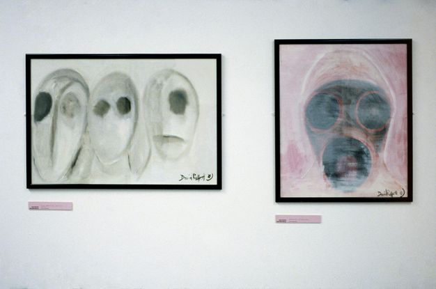 Installation photograph. Two framed artworks are hung on white gallery walls. The artwork on the left is black and white and contains three oval, head-like shapes with large black circular eyes, while the coloured artwork on the right features a mask-like form against a lilac background.
