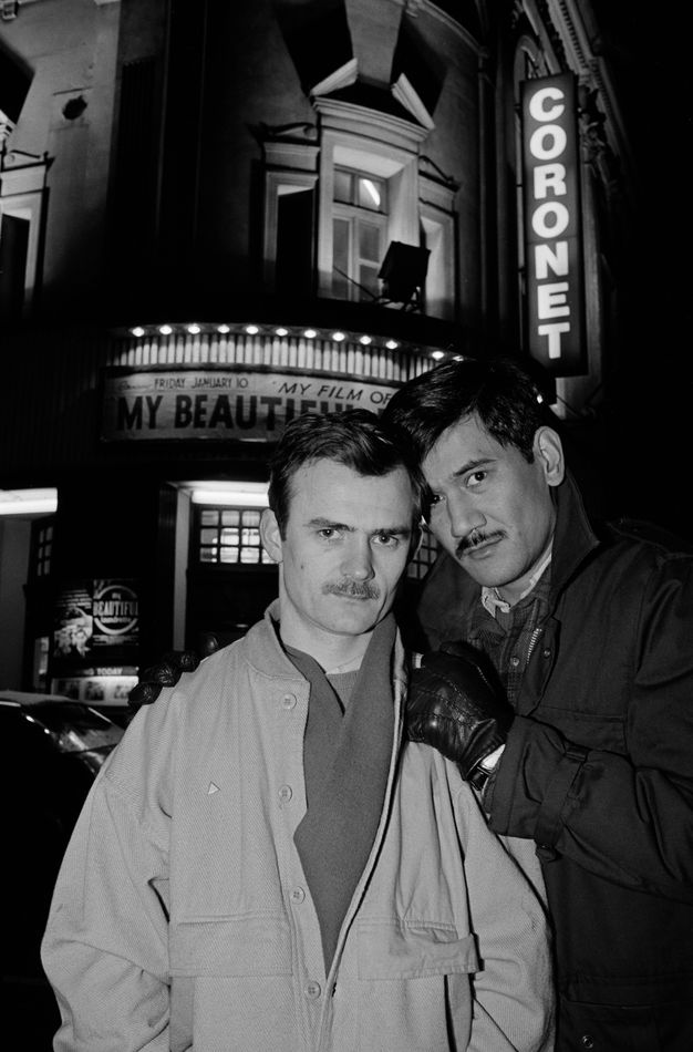 Black and white photograph. Two men stand closely together against the backdrop of a city street. Both wear winter coats and scarves, while one wears leather gloves. Behind them, a strip of vertical, illuminated text reads the word “CORONET”, while a horizontal row of bulbs lights up the signage for a cinema.
