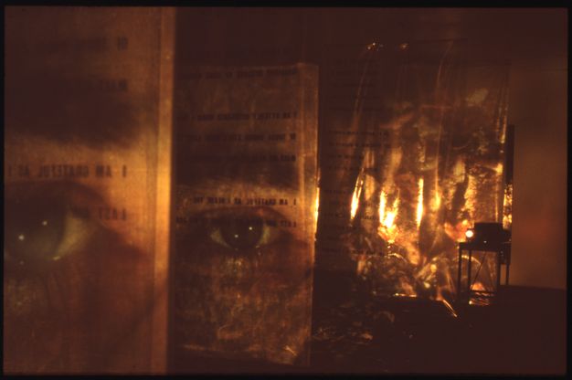 Installation photograph. In a darkened room, a series of suspended, parallel fabric sheets are projected on to with colour film. On two of the sheets, an eye is surrounded by sections of text. The projector can be seen resting on a stand in the background.