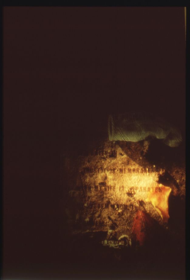 Installation photograph. This installation photograph shows a still image from a colour film projection. An almost entirely black background has a small passage of golden light in the lower right corner, that illuminates a section of text surrounded by objects.