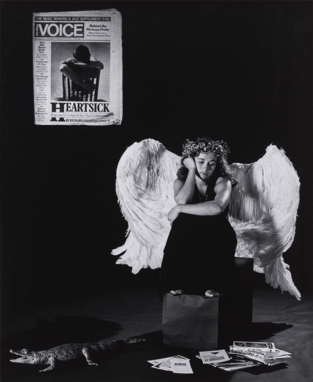Black and white photograph. A young woman wearing large, white feathered angel wings, a black dress, and a floral crown, is crouched on a stool. She rests one arm across her knee and the other on the side of her face, while looking at the ground. On the floor are a pile of magazines and leaflets, along with a toy plastic alligator, while a copy of The Village Voice is projected on the wall.