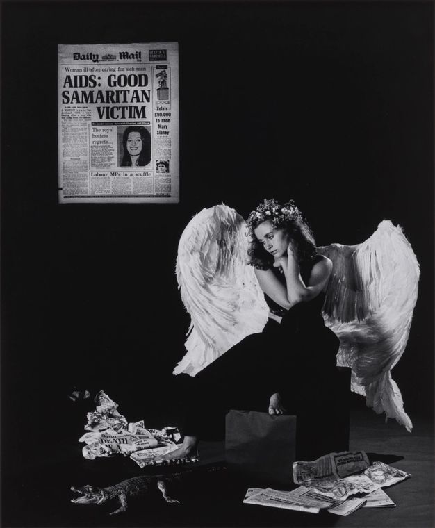 Black and white photograph. A young woman wears large white angel wings and sits on a stool with her head turned to the floor. Around her is a crumpled pile of newspapers, and a plastic toy alligator. On the left, a copy of the Daily Mail with the headline, “AIDS: GOOD SAMARITAN VICTIM” is projected on the wall.