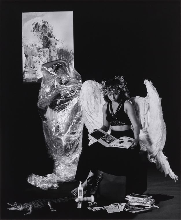 Black and white photograph. A woman wearing large, white angel wings sits intimately on the shoulders of another, who is wearing mesh wings adorned with metal objects. Magazines, a sex toy and a toy alligator are strewn on the floor below, while an artwork depicting three angels is projected onto the wall.