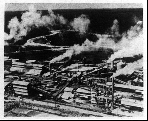 Black and white photograph. An elevated view looking over an industrial factory site. Smoke billows out of tall chimneys, forming white puffs that drift up into the sky. In the distance, a meandering road can be seen snaking into the dark land beyond.